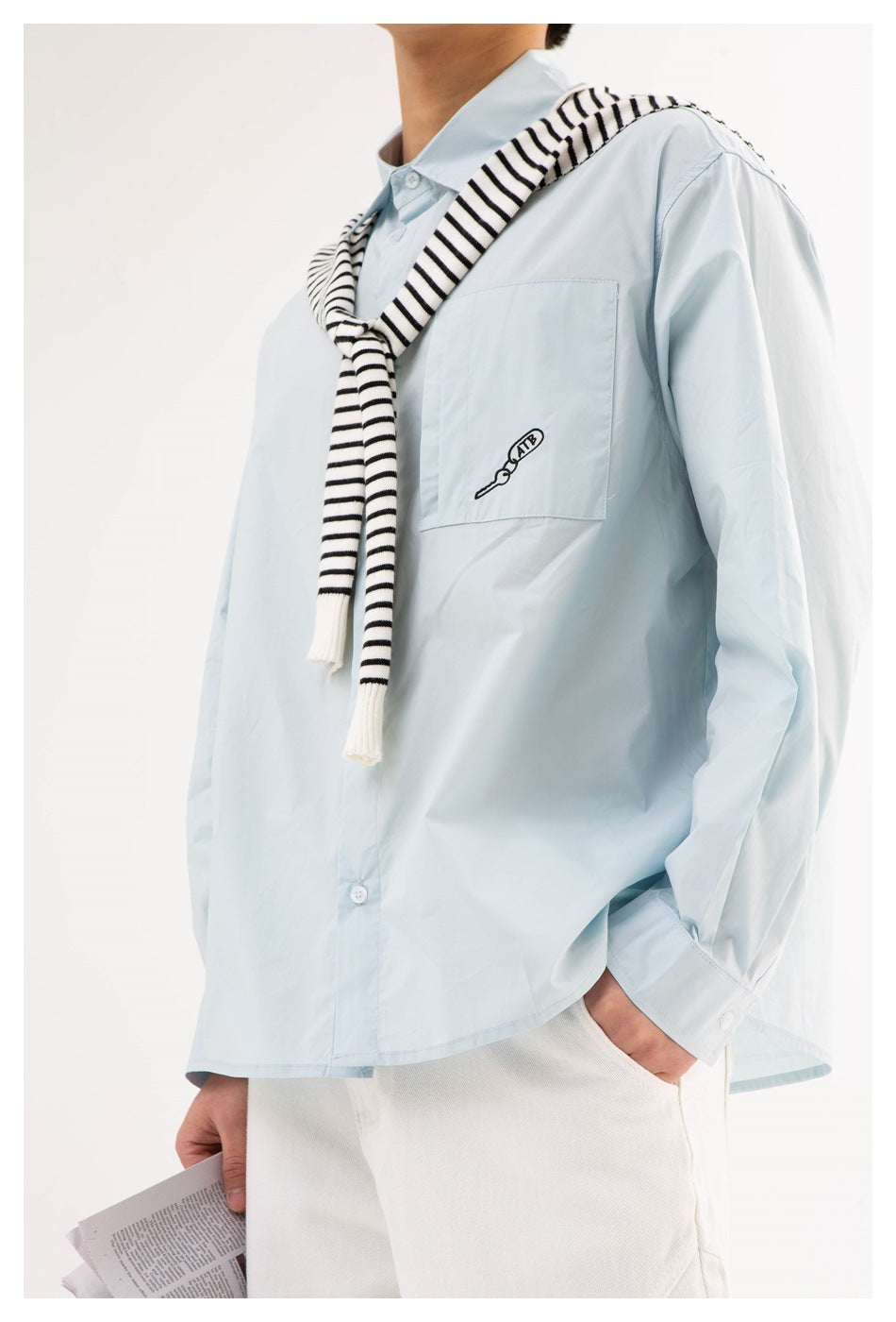 Close view of a model wearing a pale blue button-up shirt with a chest pocket and a black-and-white striped sweater draped