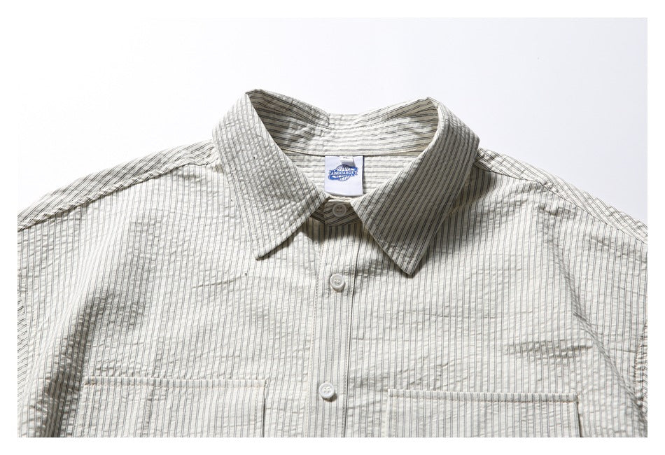 Close view of a cream button-up shirt with fine dark vertical stripes, a pointed collar, white buttons and two chest pockets.
