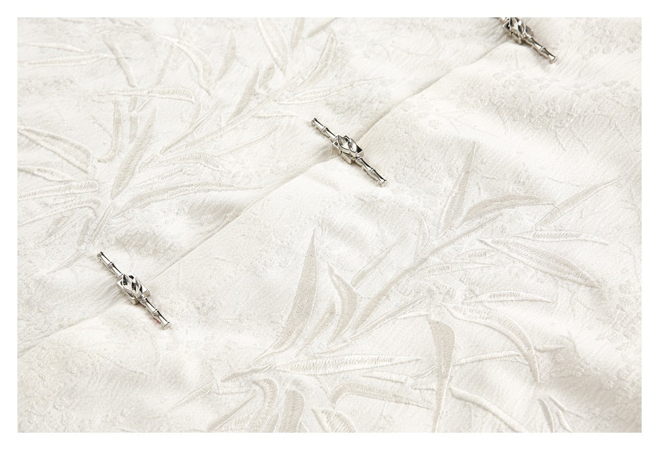 Ivory fabric close-up showing raised foliage embroidery and three silver frog-style fasteners.