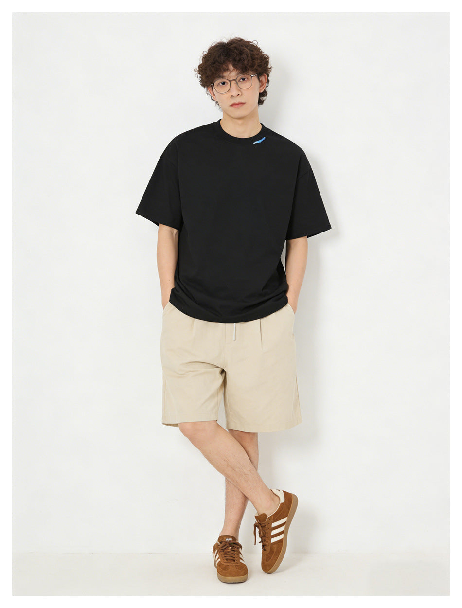 Man wearing black relaxed t-shirt with blue collar embroidery, beige shorts and brown sneakers, hands in pockets