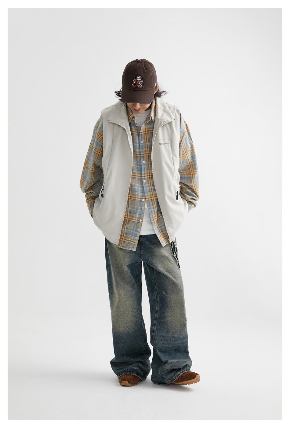 Front view of a model wearing a light gray zip-front vest over a blue and tan plaid shirt and loose jeans.