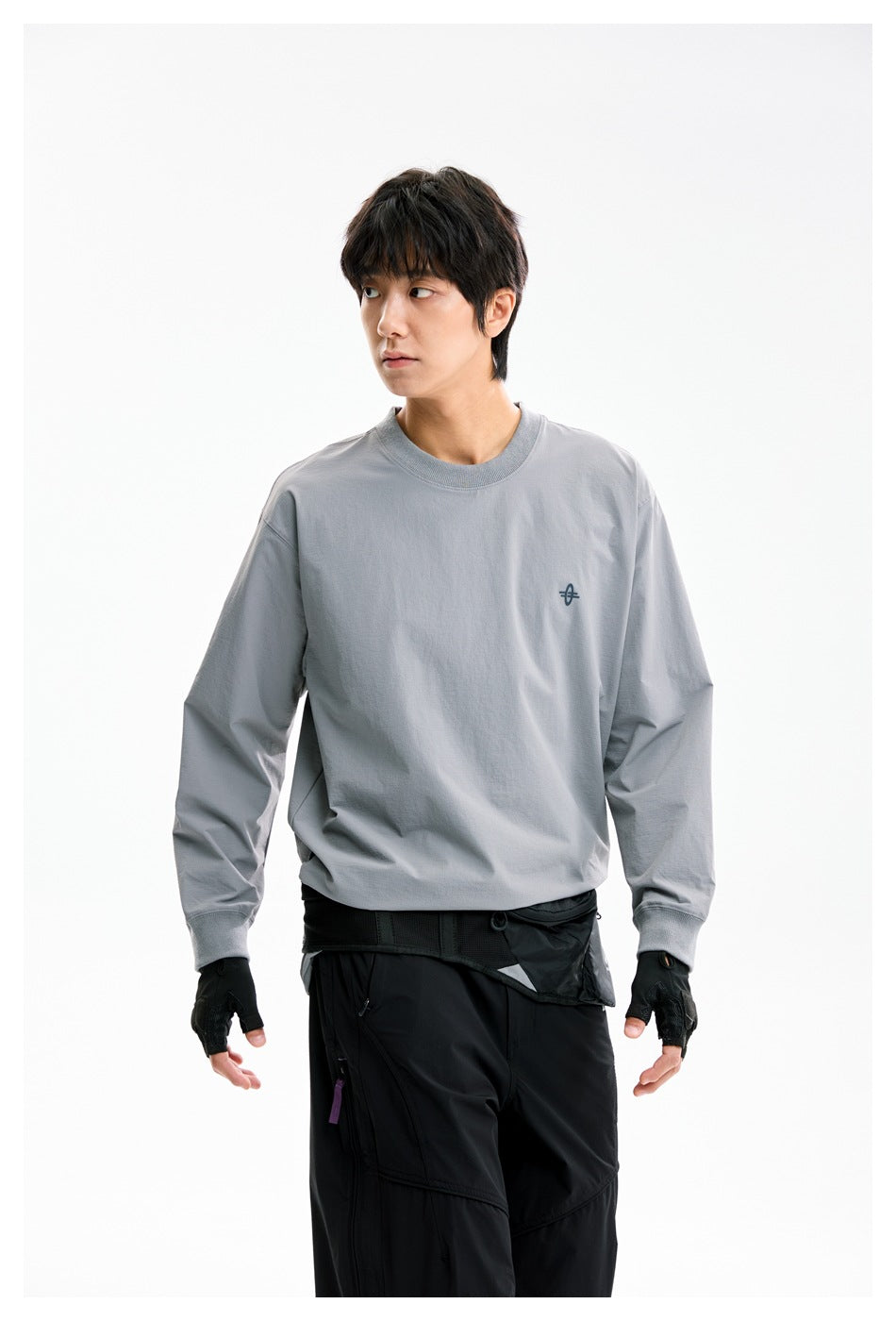 Model wearing a gray long-sleeve crewneck with a small chest emblem and black pants, looking to the side.