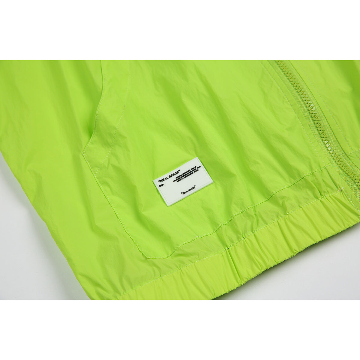 Close-up of a lime green jacket showing a zipper, stitched panel, gathered hem, and a small REAL SPACE label.