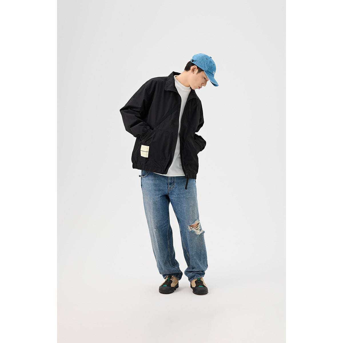 A model wears a black zip jacket open over a white shirt, with blue jeans and a blue cap.