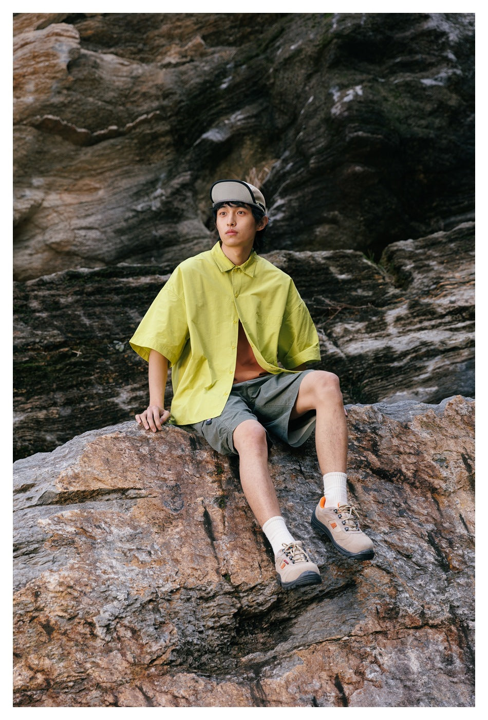 A seated man wears a lime-yellow short-sleeve button-up shirt and gray cargo shorts on a rocky slope.
