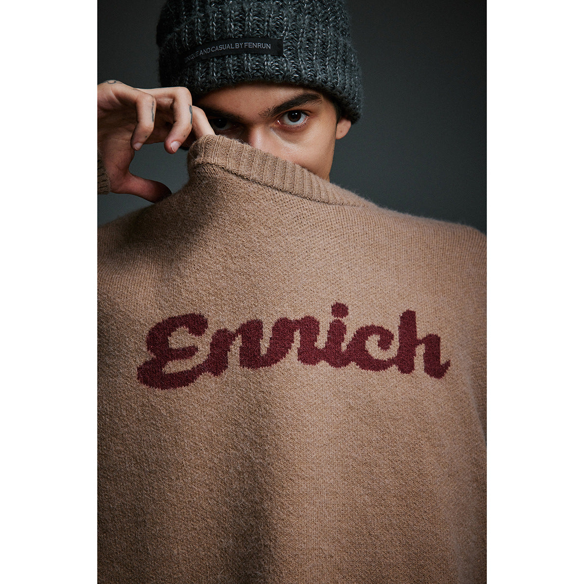 Close-up of a model in a tan sweater with burgundy script lettering and a dark ribbed beanie, holding the collar over his