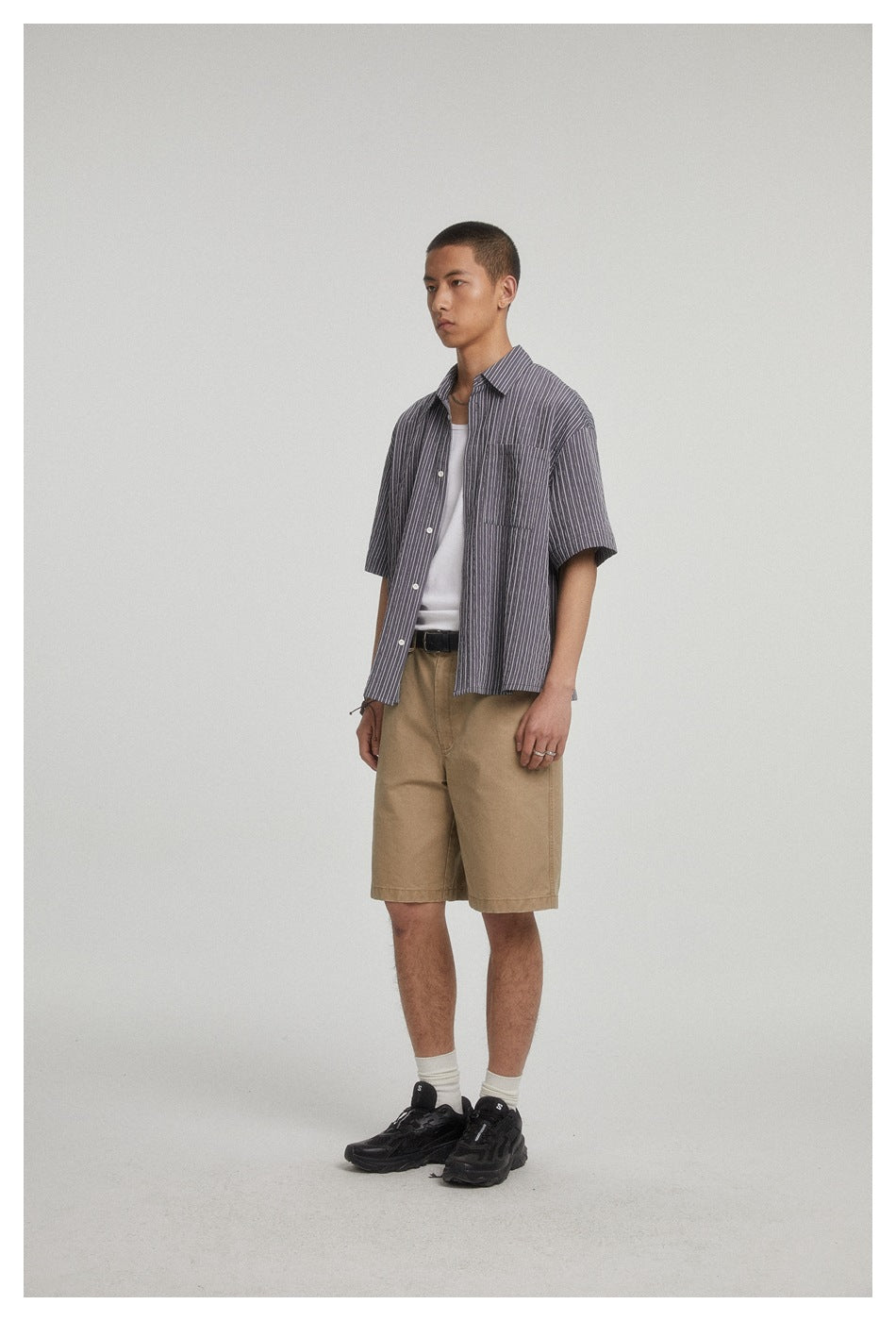 Model wearing an open charcoal gray pinstriped short-sleeve shirt over a white top with tan shorts.