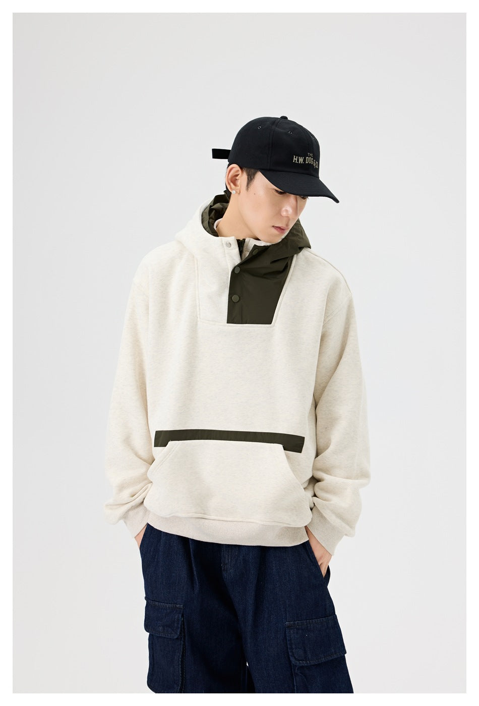Model looking down in oatmeal color-block hoodie with olive panels, hands in pockets, black cap and dark denim cargo pants