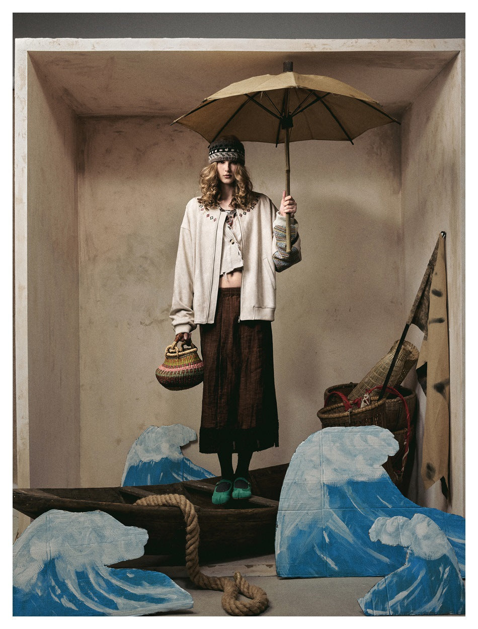 Model in a beige zip jacket with neckline embroidery and patterned sleeve trim, holding an umbrella in a styled set