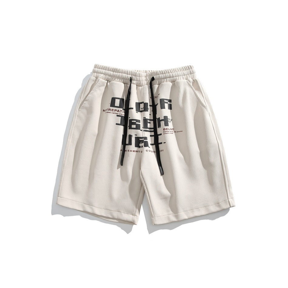 Khaki drawstring shorts with large black pixel-style lettering graphic on the front, flat lay