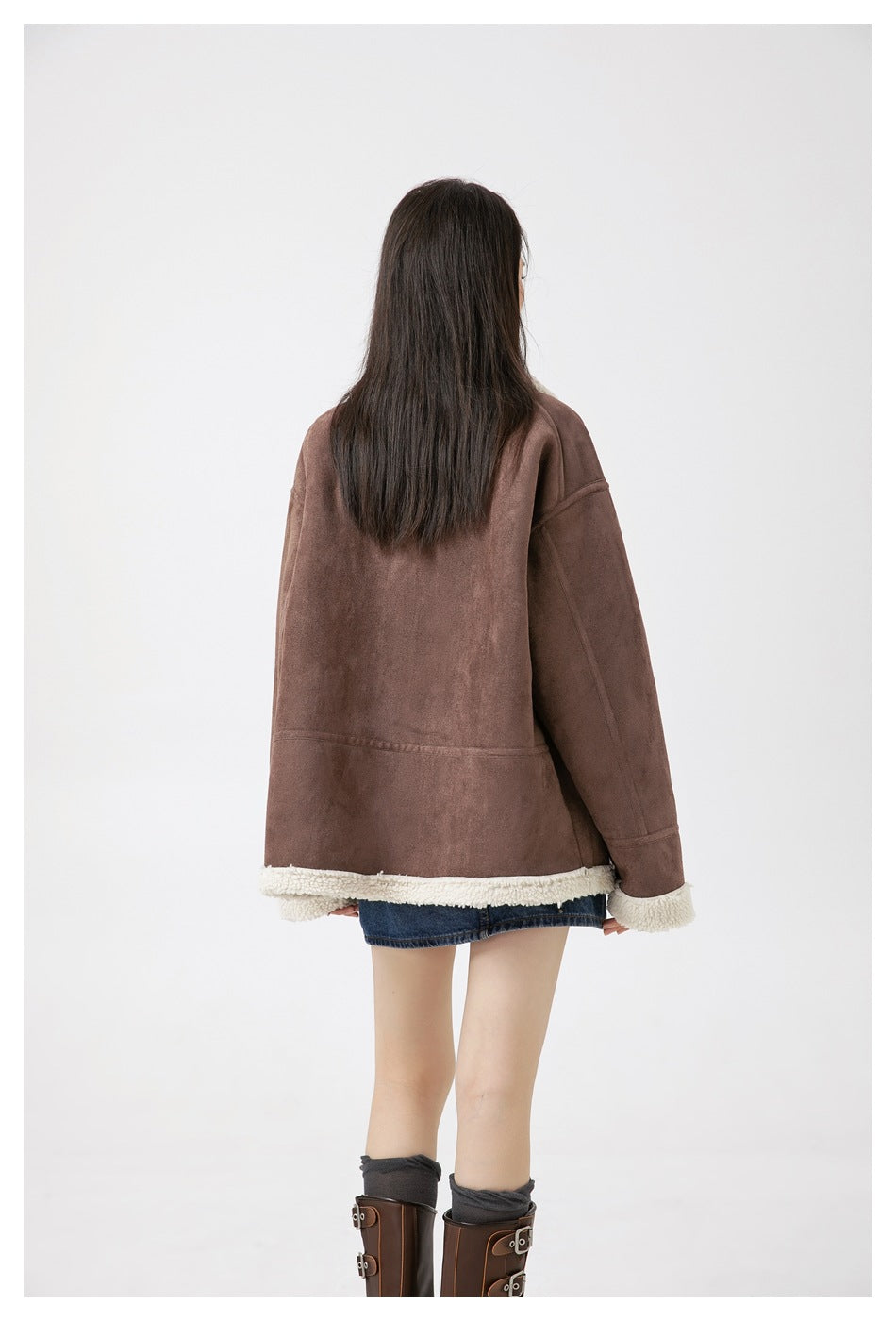 Rear view of a model wearing an oversized brown jacket with cream sherpa trim, denim shorts and tall boots.