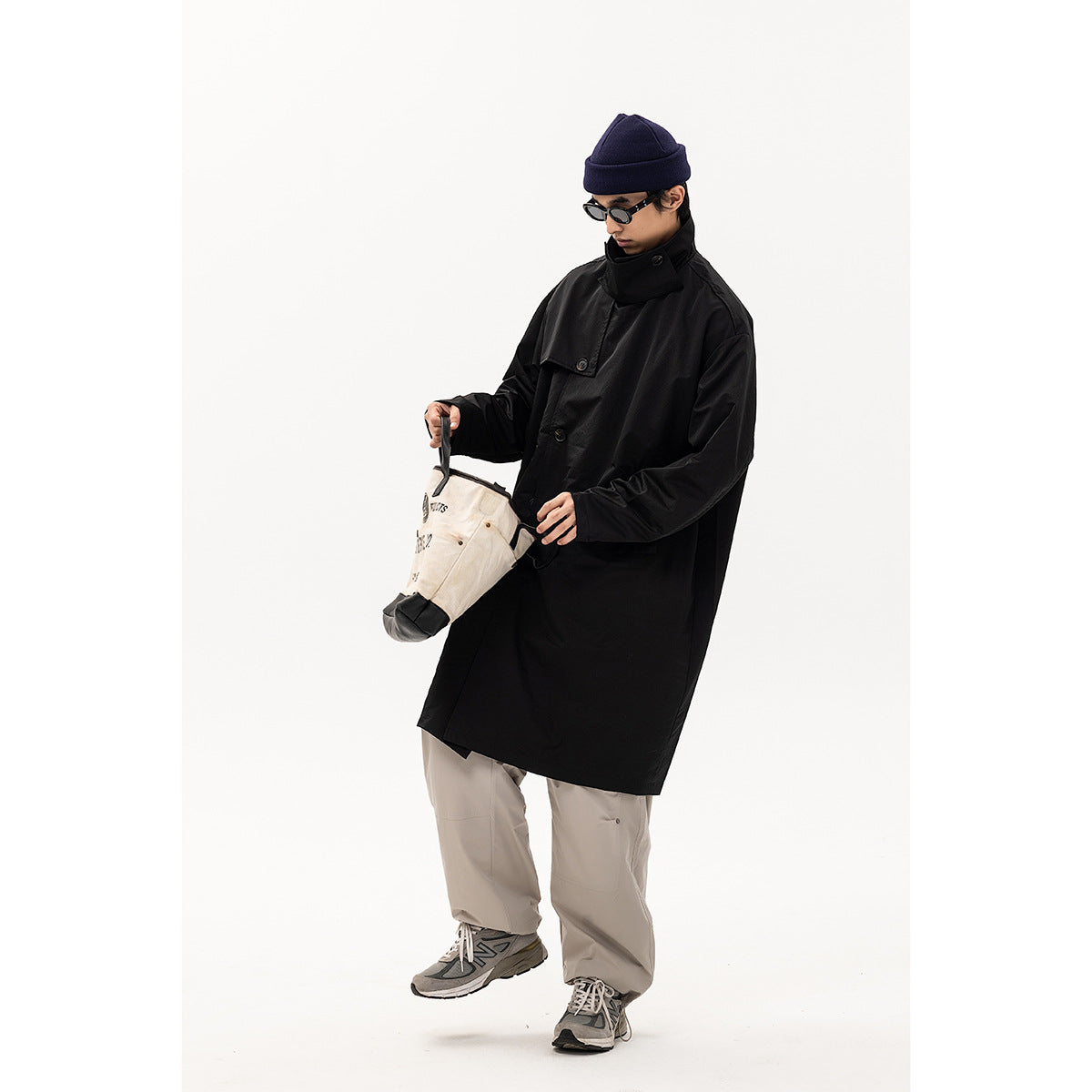 Model in a long black coat with a high buttoned collar, navy beanie and gray trousers, holding a cream bag.