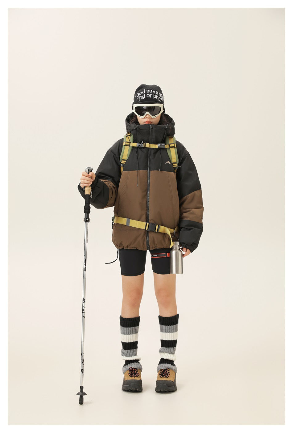 Full-length model wearing a black-and-brown hooded jacket with hiking gear and striped leg warmers.