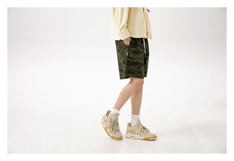 Side view of a model wearing dark green camouflage shorts with a cream top, white socks and tan sneakers.