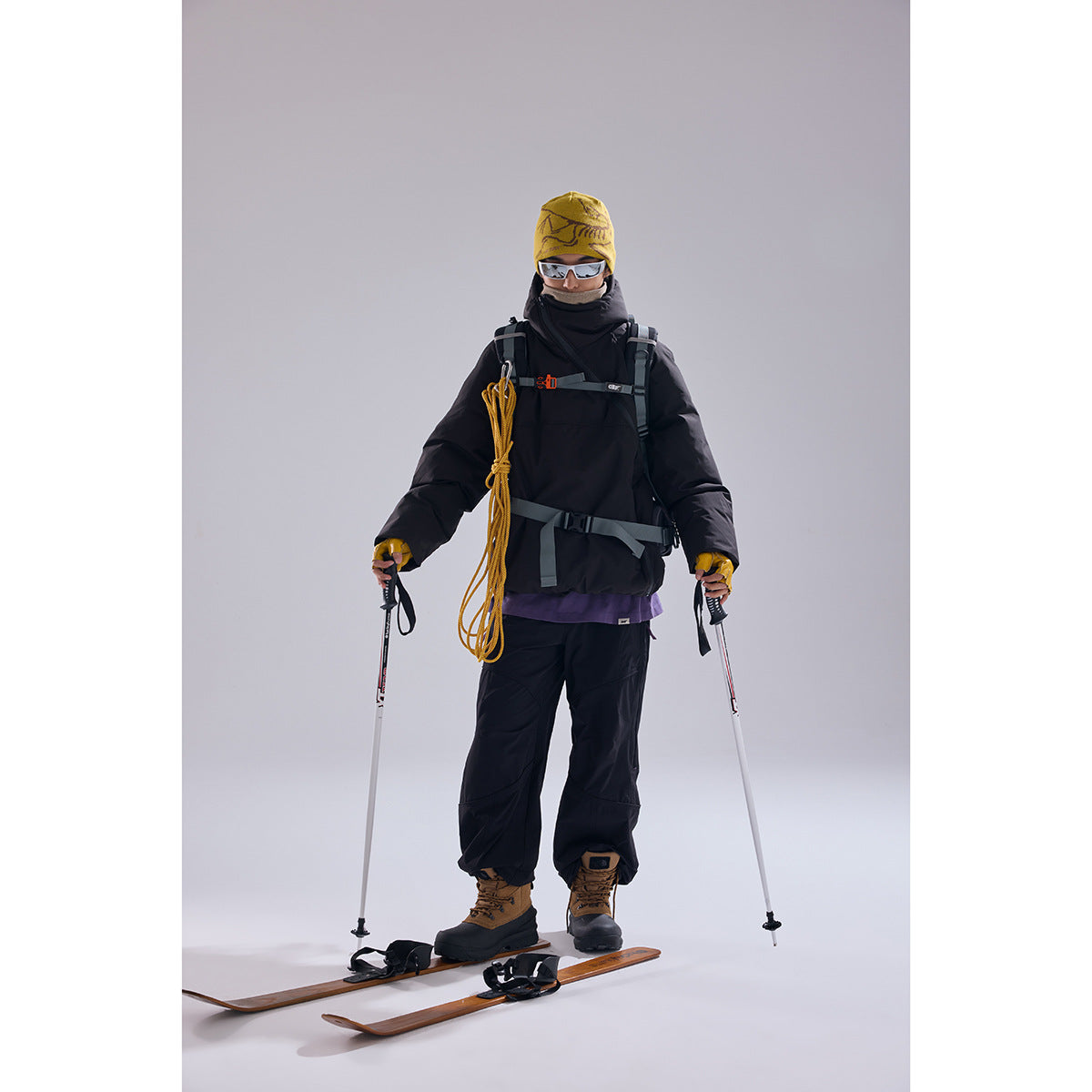 Model in a black hooded winter jacket and pants with a yellow beanie, ski poles, and skis.