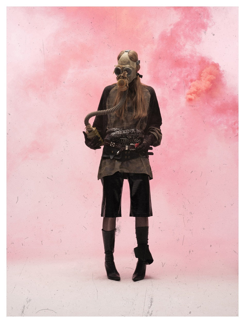 Model in a gas mask and belts wearing a camouflage top with dark sleeves and black shorts against pink smoke