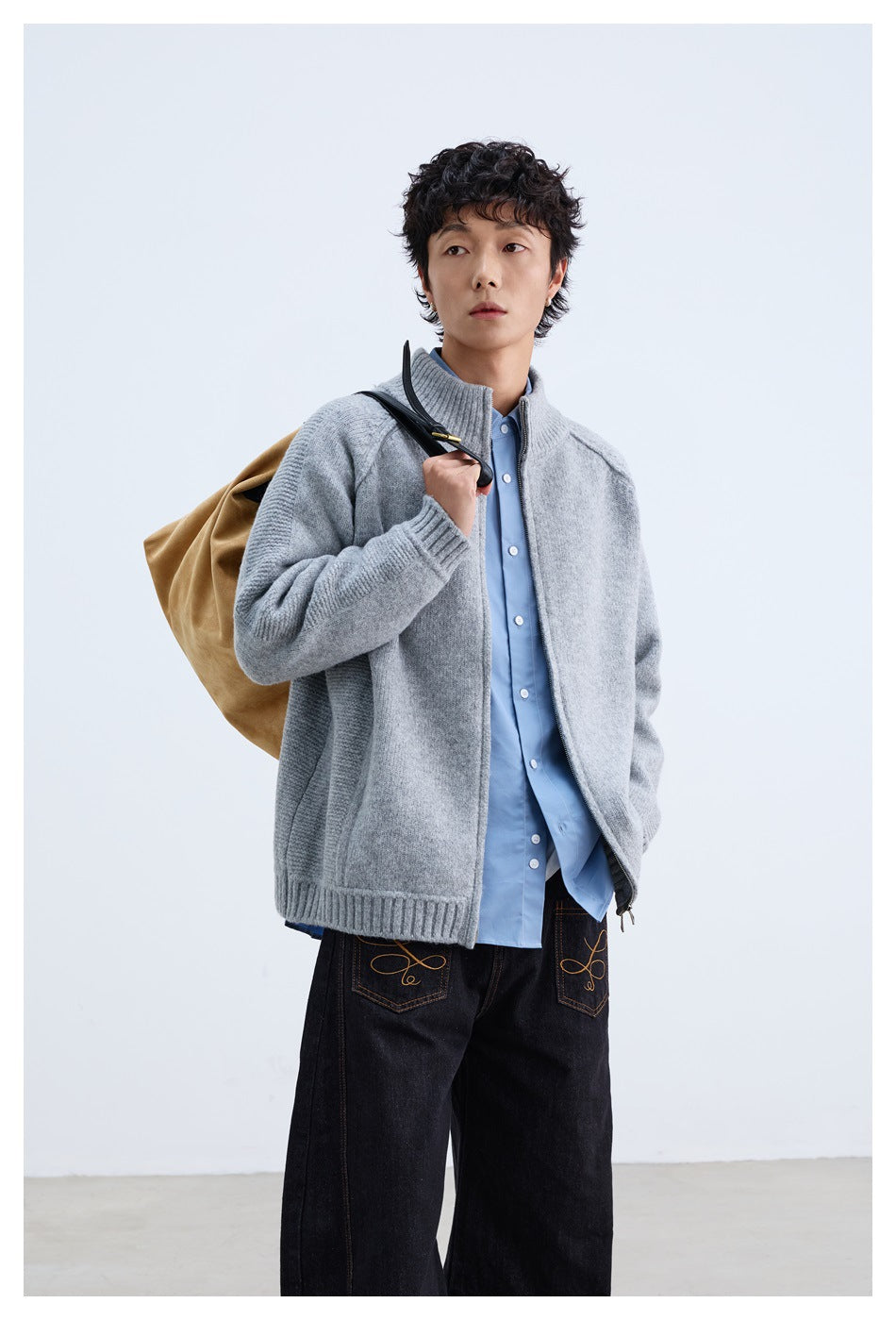 Model wearing an open light gray zip-front cardigan over a blue shirt, carrying a tan bag.