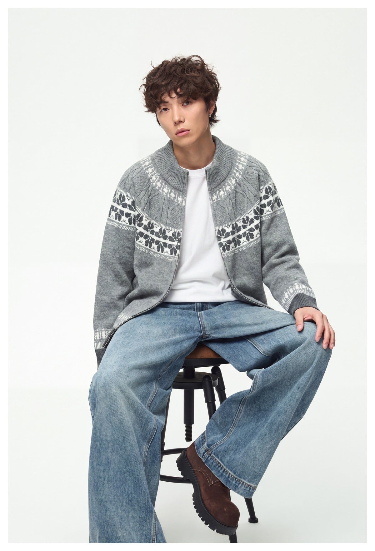 Seated model wearing open light gray snowflake-pattern zip knit sweater over a white tee with light blue jeans
