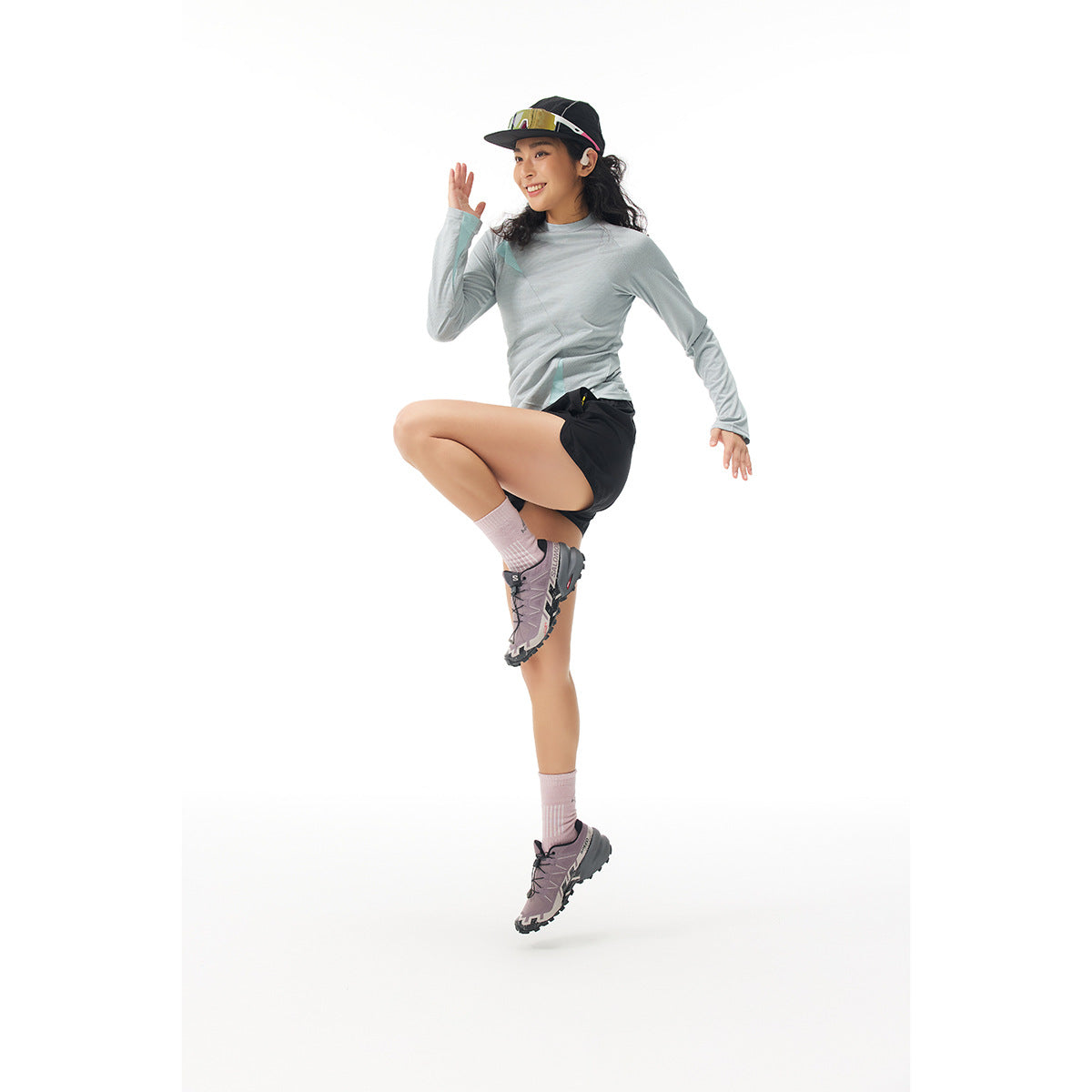 Model in a light gray-green long-sleeve top and black shorts, posing mid-jump in a cap.
