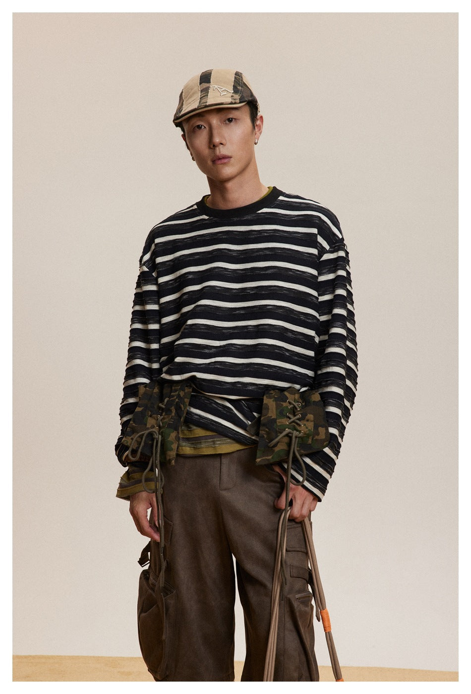 Model wearing a loose black-and-cream striped long-sleeve shirt, shown from the front with brown trousers and a tan cap.