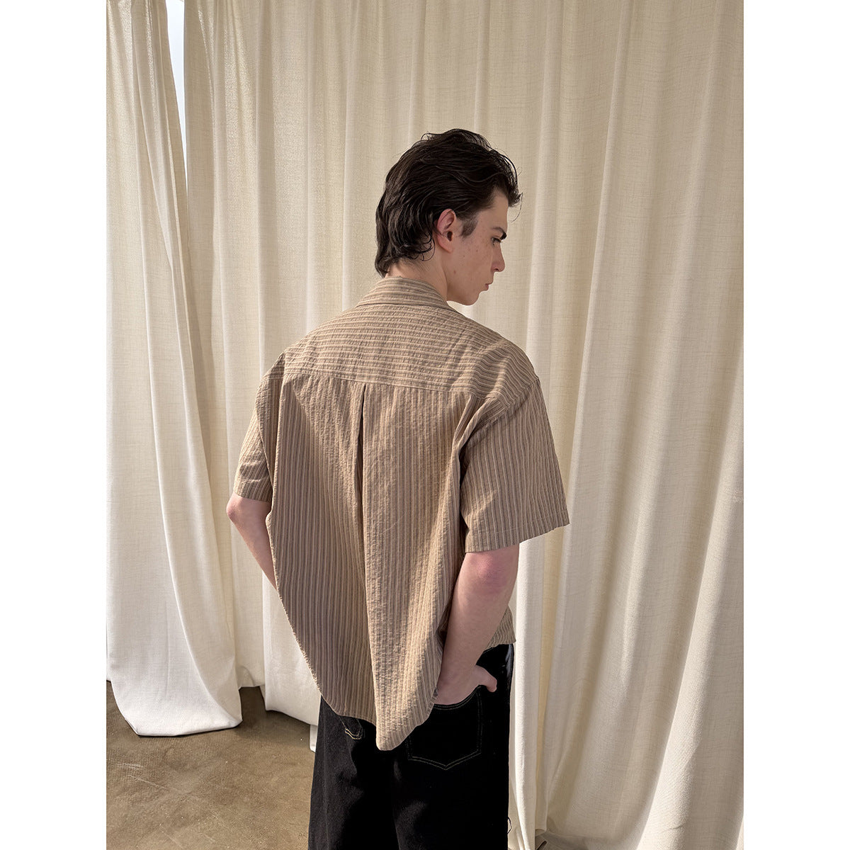 Rear view of a model wearing a tan vertically striped short-sleeve shirt with a back yoke and center pleat.