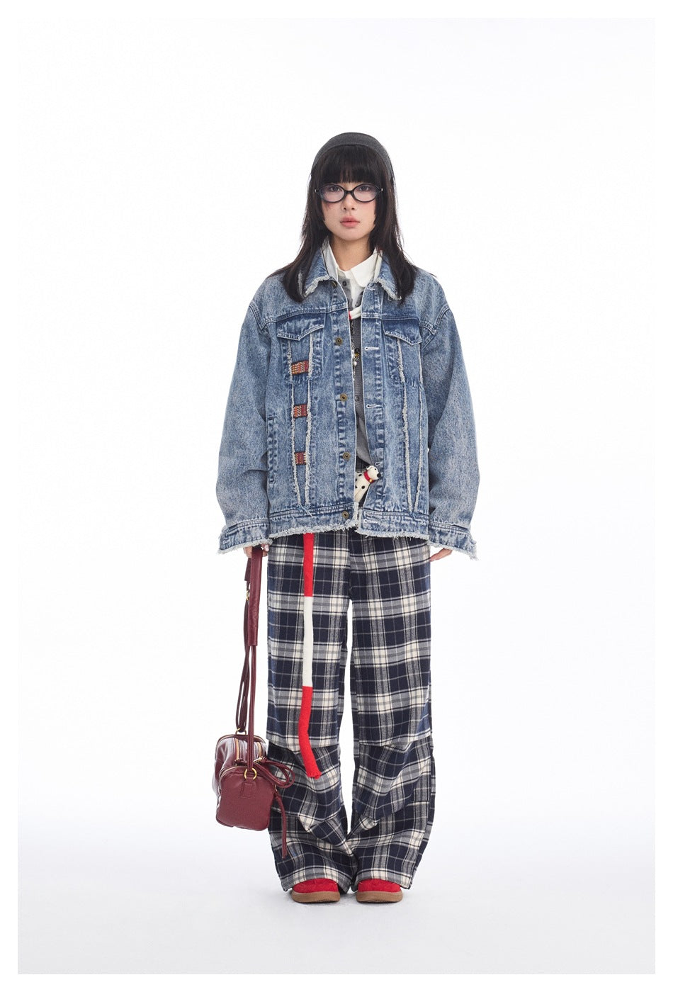 Model wearing a blue denim jacket with decorative patches and loose black-and-white plaid trousers against a white