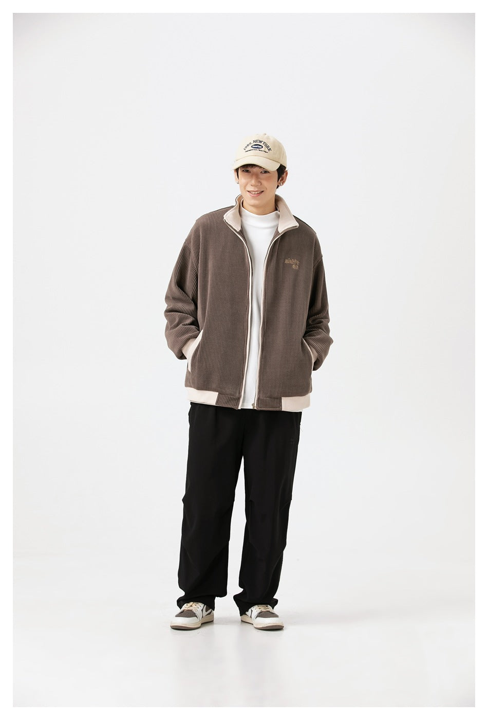 Full-length view of model in brown ribbed zip jacket with cream trim, white top, black pants and sneakers