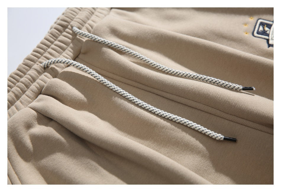Close-up of beige jogger pants showing two braided drawstrings with dark metal tips and part of an embroidered patch.