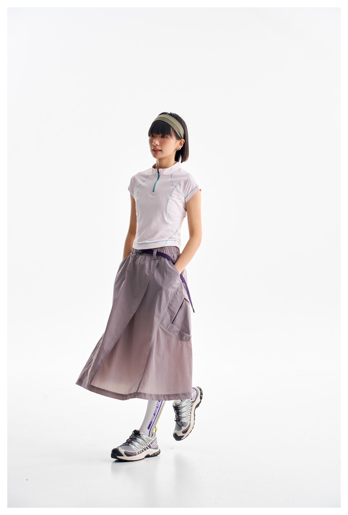 Model in a white zip-neck top with teal seam accents and a gray-lilac wrap skirt, shown in a walking pose.