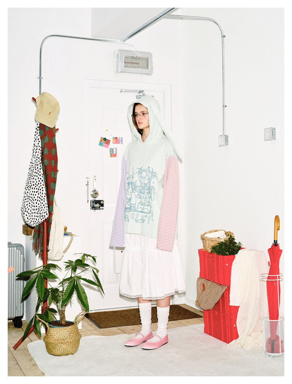 A person models a pale mint bunny-ear hoodie with pink and lavender striped sleeves and a blue cartoon chest graphic, with a
