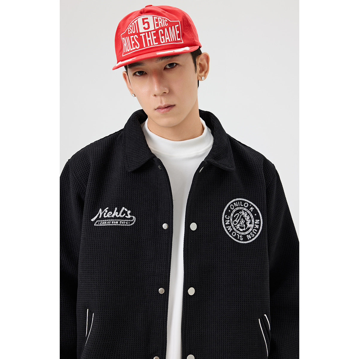 Close-up of a model wearing a black snap-front jacket with white chest embroidery and a red text cap.