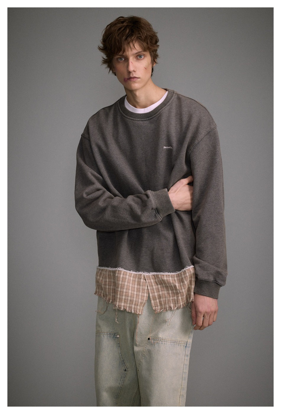 Man modeling a charcoal oversized sweatshirt with a frayed plaid hem, layered over loose pale jeans.