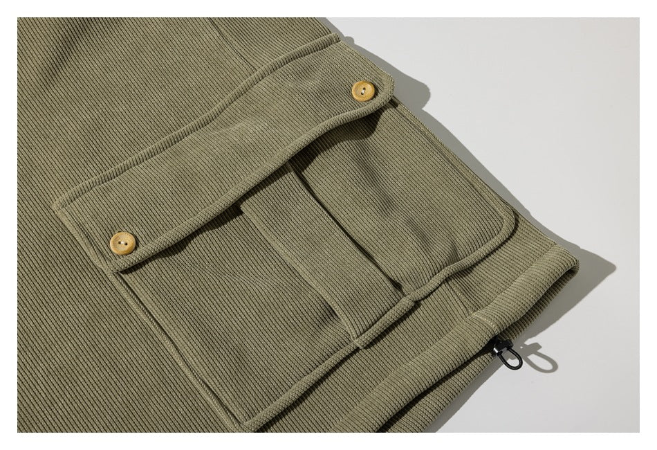 Close-up of an olive ribbed garment pocket flap with two tan buttons and a black loop at the edge.