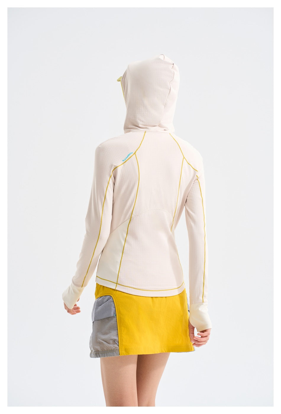 Rear view of a model wearing a pale hooded jacket with yellow seam piping and a color-block yellow and gray skirt.