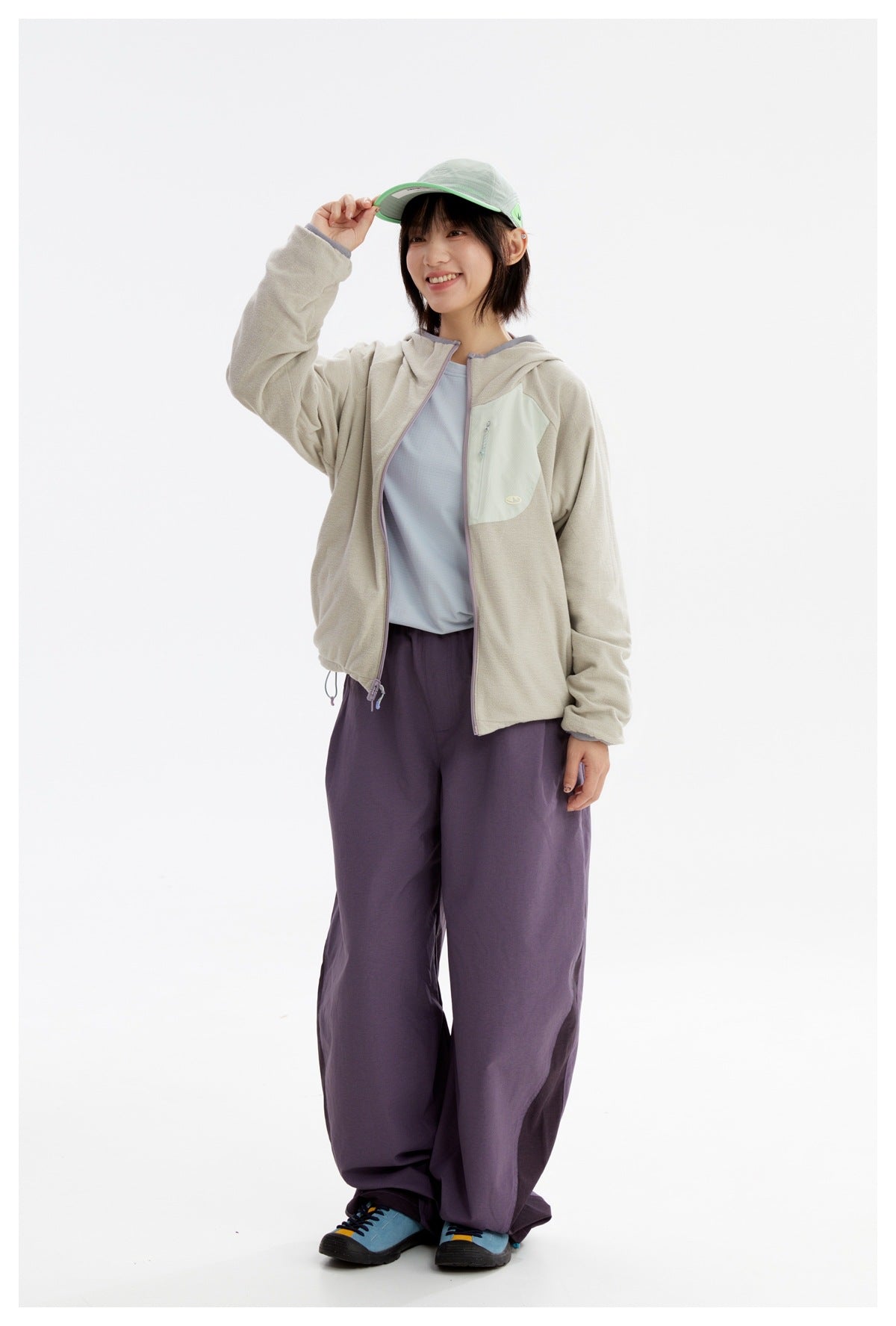 Model wearing a light gray-beige hooded zip jacket with a pale mint chest pocket and dark purple trousers.