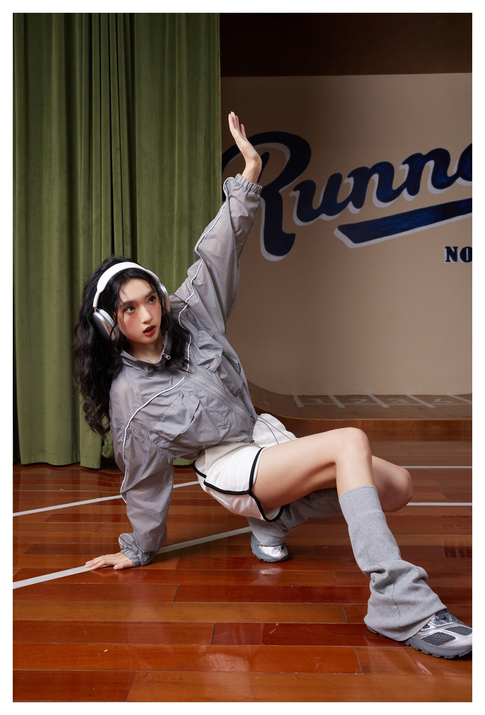 Model wearing a light gray cropped zip jacket with white shorts and gray leg warmers, seated on a court floor.