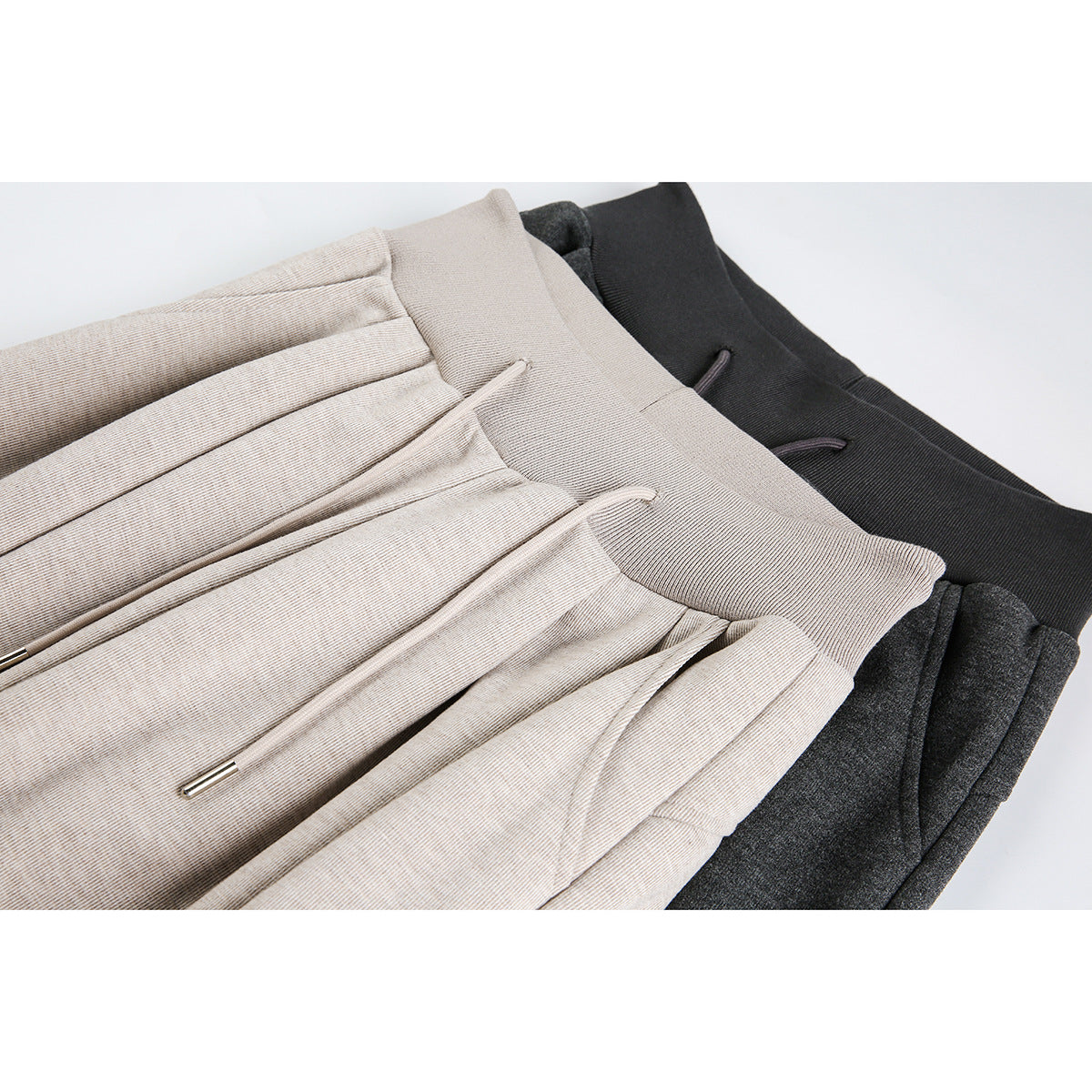 Close-up of folded wide-leg drawstring pants in light beige, charcoal gray, and black, showing pleats and waistbands.
