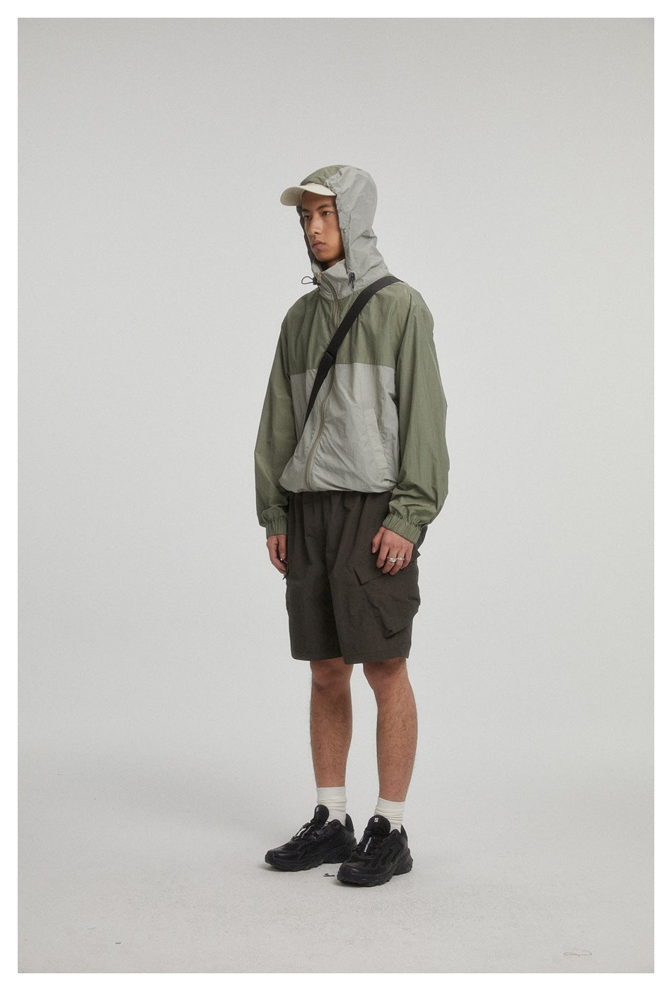 Model wearing a hooded two-tone olive and light gray zip jacket with dark cargo shorts.