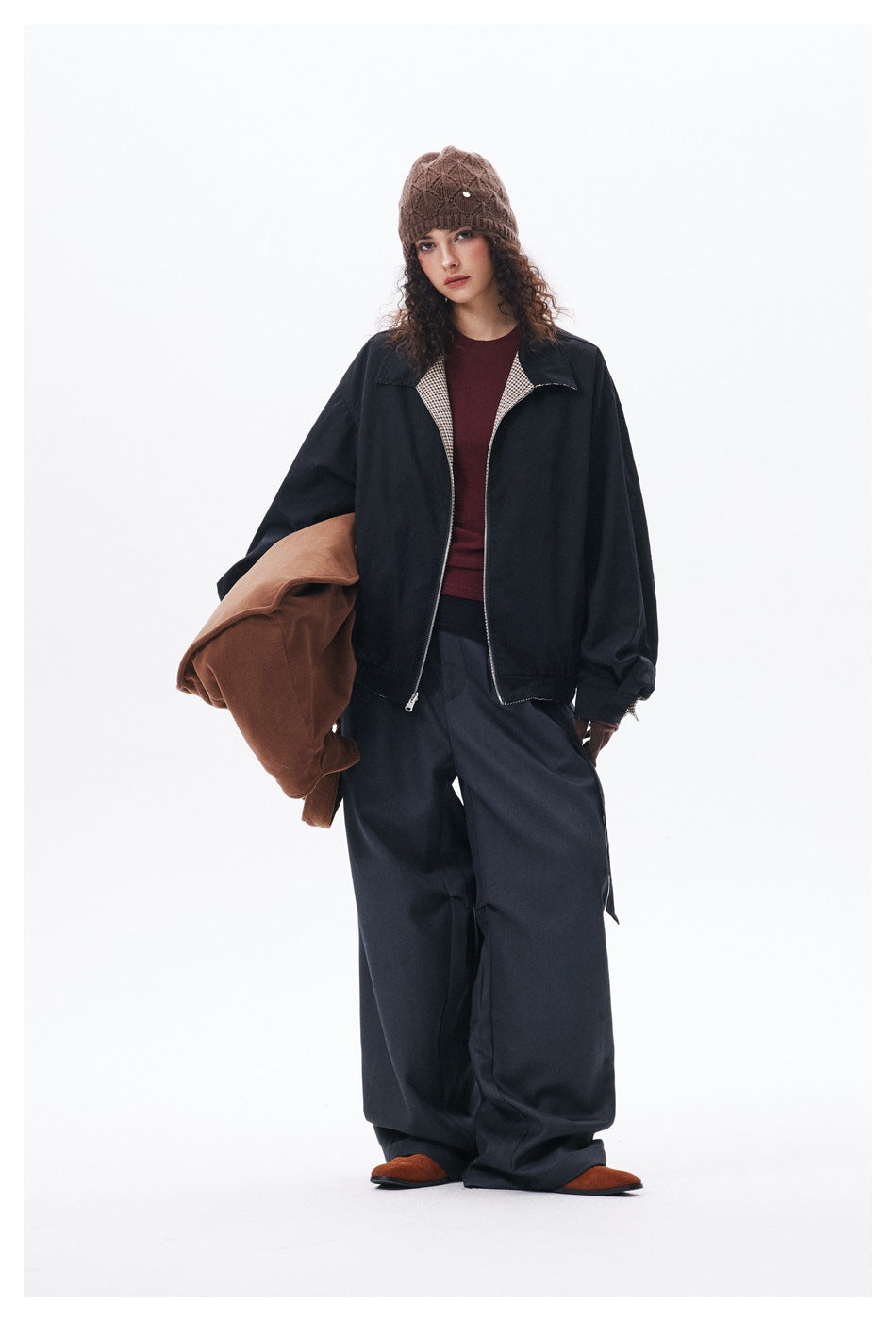 Full-length model wearing a black zip jacket with checked lining, burgundy top, loose dark trousers and a brown knit beanie.