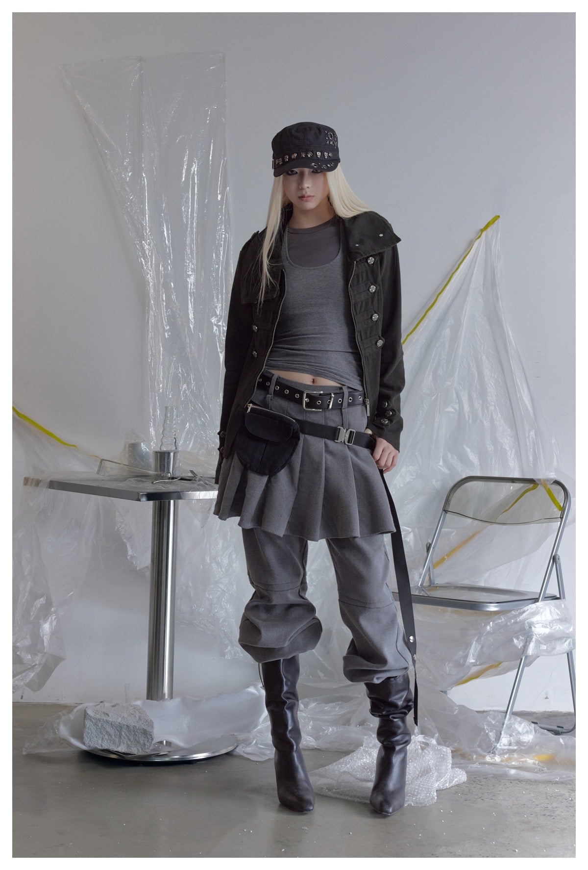 Model wearing the black jacket open over a gray tank top, gray pleated skirt, gray pants, black belt, and boots