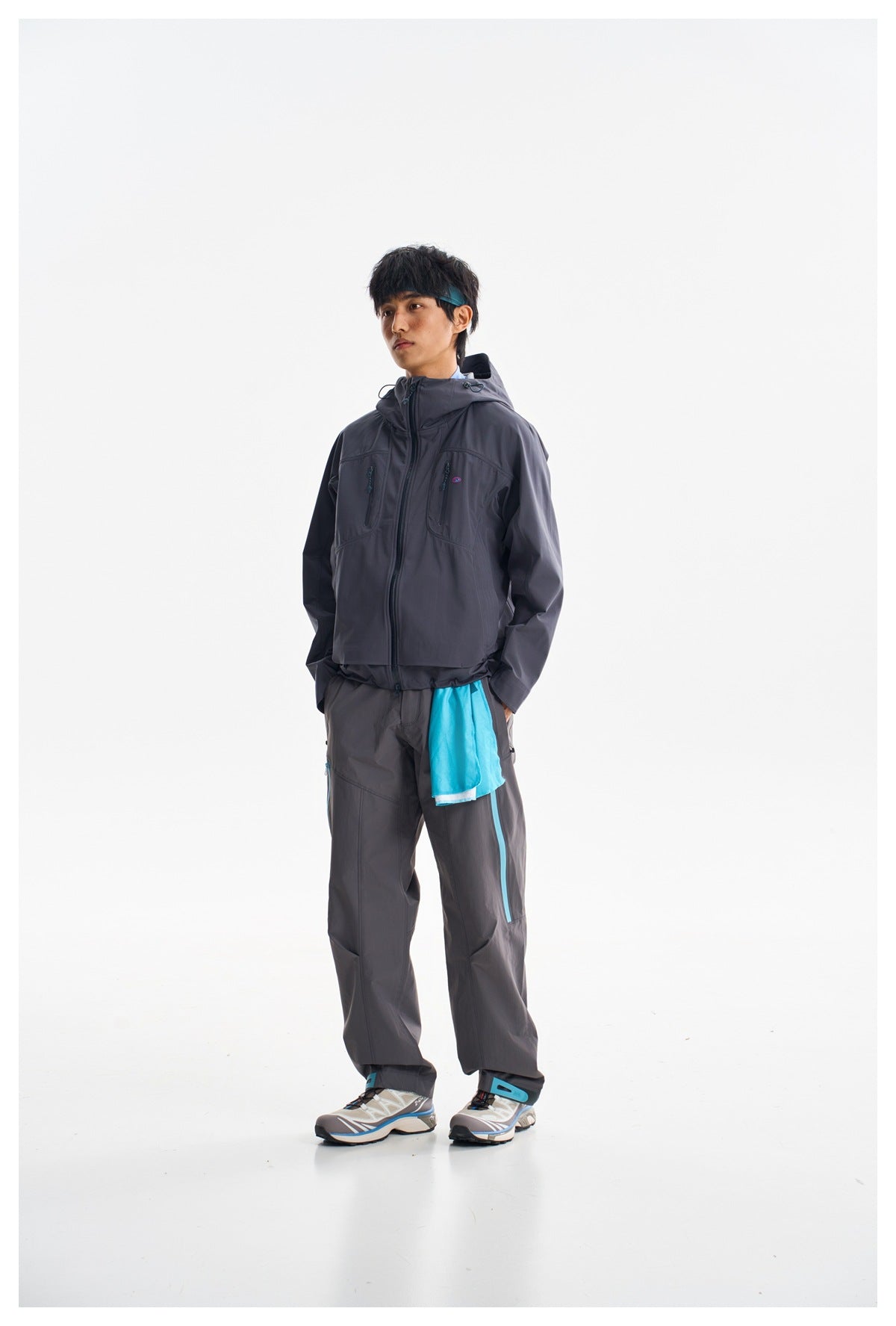 Model wearing a charcoal hooded jacket and matching trousers with bright turquoise accents, shown full length.