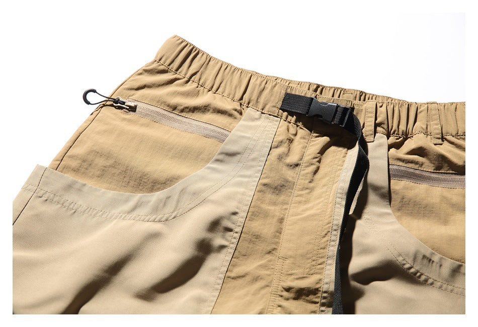 Close-up of tan utility shorts showing the elastic waistband, black buckle belt, zipper pockets, and beige side pocket.