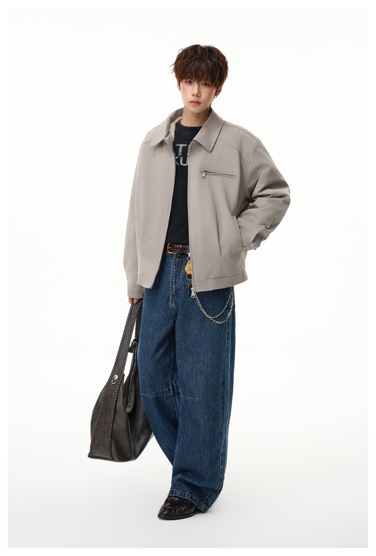 Full-length view of a model in a light taupe zip jacket and loose dark blue jeans, carrying a dark tote.