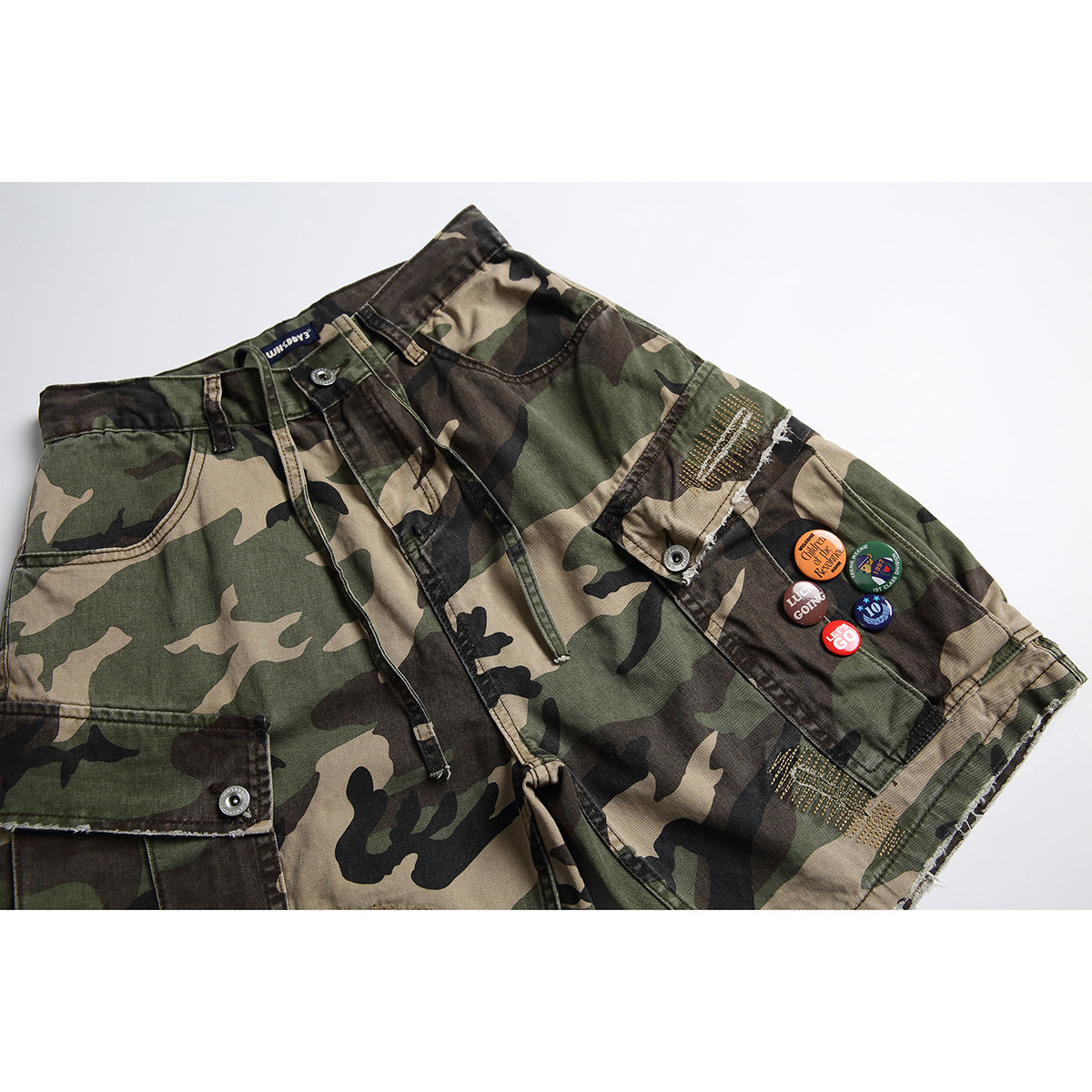 Front view of olive, tan, and black camouflage cargo shorts with frayed hems, gold stitching, and decorative pins.