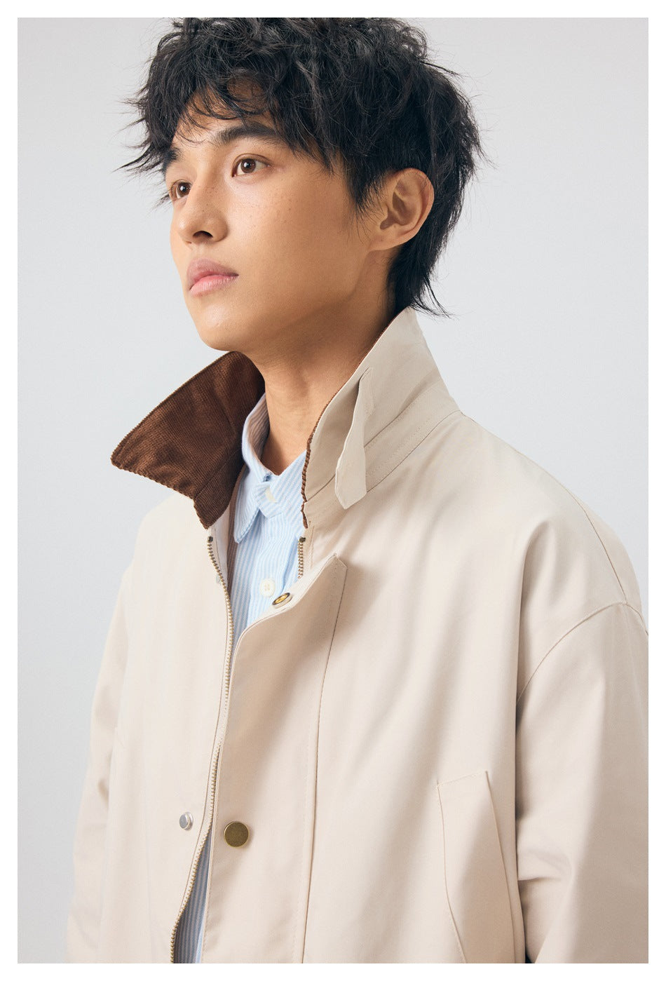 Close-up of a model in a beige coat, highlighting the raised collar, brown underside, zipper, and snaps.