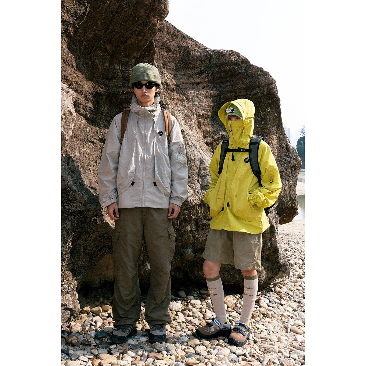 Two people wear light gray and bright yellow hooded outdoor jackets beside a rocky shore.