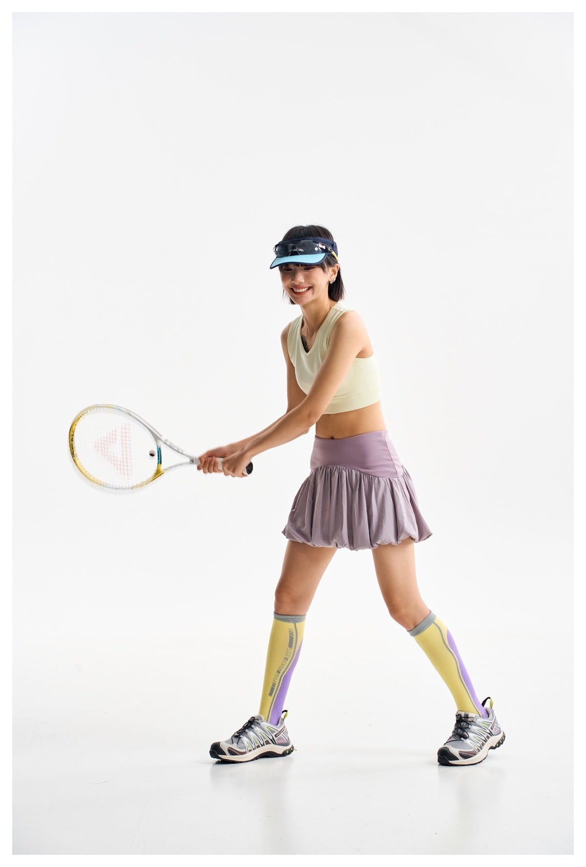 Model in a pale yellow crop tank and lilac bubble-hem skirt swings a racket in a tennis pose.
