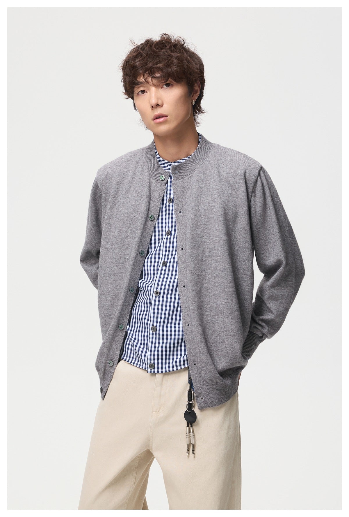 Model wearing an open gray button-front cardigan over a blue-and-white checked shirt and cream trousers.