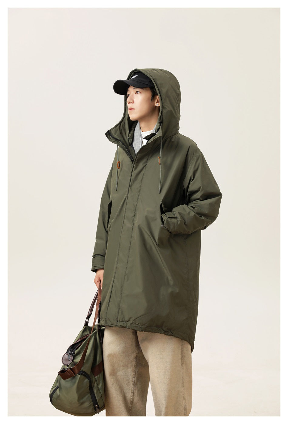 Model wearing a dark olive hooded parka with orange cord toggles, shown at an angle with a bag.