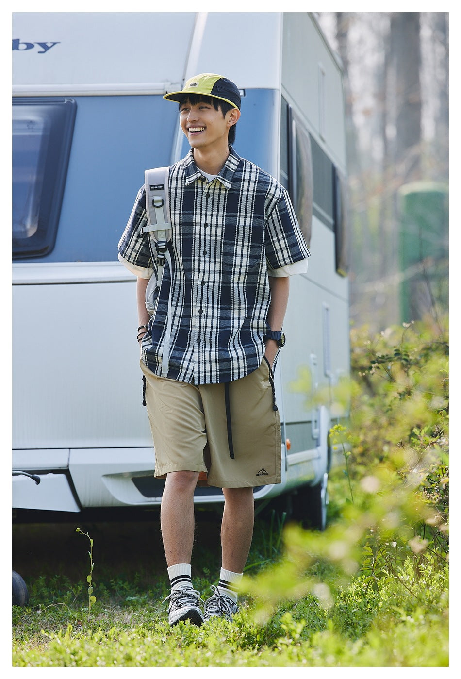 Smiling man walking outdoors in navy plaid short-sleeve shirt, khaki shorts, cap and backpack